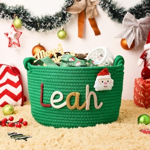 May include: A green woven basket with the name "Leah" in colorful letters and a Santa Claus applique. The basket is filled with green ribbon and other Christmas decorations. The basket is sitting on a fluffy, light brown rug.