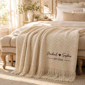 Può includere: Una coperta in maglia testurizzata color crema con lunghe frange sul bordo inferiore. La coperta è personalizzata con un testo marrone scuro che riporta Michael, un simbolo di cuore, Sophia e il testo EST.2026 tra due linee orizzontali.