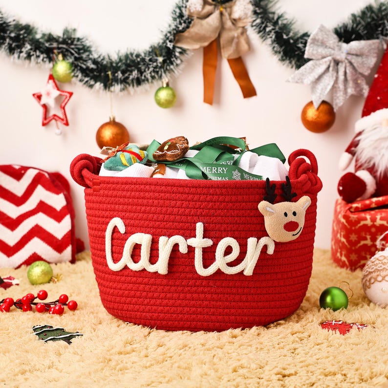 May include: A red woven basket with the name "Carter" in white script. The basket is decorated with a reindeer and filled with gifts and ribbons. Christmas ornaments and decorations surround the basket.