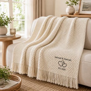 May include: A cream-coloured textured knit throw blanket with fringe trim on the edges. The blanket features black embroidered text that reads Samuel & Hannah, a graphic of two interlocking hearts, and the text EST. 2026 printed below the hearts.