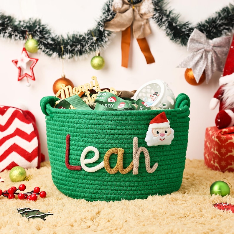 May include: A green woven basket with the name "Leah" in colorful letters and a Santa Claus applique. The basket is filled with green ribbon and other Christmas decorations. The basket is sitting on a fluffy, light brown rug.