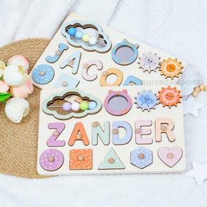 May include: Two wooden name puzzles with colourful letters and shapes. One puzzle spells "JACOB" and the other spells "ZANDER". Each puzzle has a cloud-shaped bead maze and animal-themed mirrors and gears.