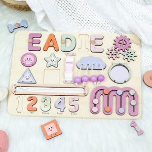 May include: A wooden busy board with the name "EADIE" in colourful letters. It features various interactive elements like shapes, numbers, a zipper, and a buckle. The board is designed with pastel colours and playful illustrations.