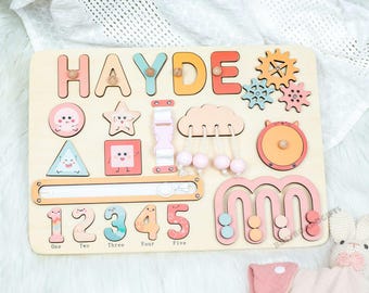May include: Wooden educational puzzle board with the name "HAYDE" in colorful letters. Features shapes, numbers, gears, and a bead maze. Includes the numbers one through five. The board is light brown with pastel-colored accents.