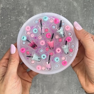 Può includere: Uno stampo in silicone trasparente con un design circolare. Lo stampo presenta più tazze piccole con un design a tè boba e piccole forme di ciambelle. Lo stampo è riempito con resina rosa, blu e bianca.