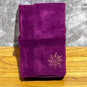 Op de afbeelding: Een dieppaarse, fluweelachtige wikkel met een flap- en riemsluiting. De wikkel heeft een goudkleurig geborduurd zonontwerp op de voorkant. Het item is waarschijnlijk ontworpen om items vast te houden en te beschermen.