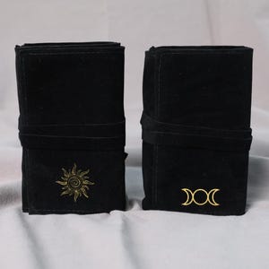 Könnte beinhalten: Zwei schwarze Wildleder-ähnliche Beutel mit umlaufenden Riemen. Einer hat ein goldenes Sonnenmuster, der andere ein goldenes Dreifachmondsymbol. Die Beutel dienen wahrscheinlich zur Aufbewahrung von Gegenständen.