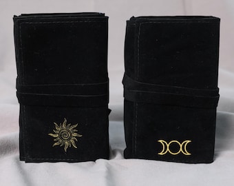 May include: Two black rectangular fabric rolls secured with matching fabric ties. The left roll features a gold embroidered sun design, while the right roll displays a gold embroidered triple moon symbol. Both items have a soft, matte texture.