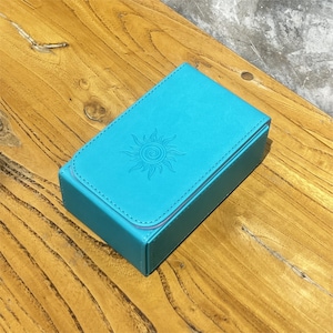 May include: A turquoise, rectangular box with a sun design embossed on the lid. The box is made of a smooth material and has a stitched border. The box is resting on a wooden surface.