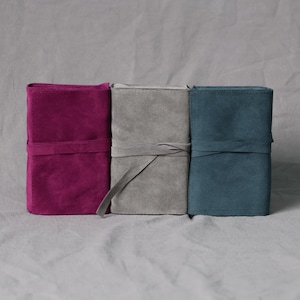 Puede incluir: Tres estuches rectangulares de ante dispuestos uno al lado del otro. El estuche de la izquierda es magenta, el central es gris claro y el de la derecha es azul verdoso. Cada estuche cuenta con un cierre de lazo de ante a juego.