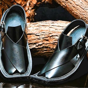 May include: A pair of black leather sandals with a distinctive design. The sandals feature a strap that crosses over the foot and a buckle closure. The soles are stitched and the sandals are displayed on a wooden surface.