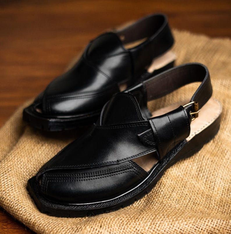 May include: A pair of black leather Peshawari sandals. These open-toe sandals feature a strap around the ankle with a buckle closure. The sandals are displayed on a tan burlap surface.