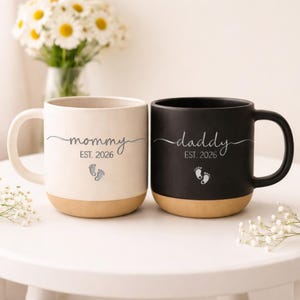 Pode incluir: Duas canecas de cerâmica, uma creme e outra preta, com bases castanhas. A caneca creme diz "mommy EST. 2026" com pegadas de bebé. A caneca preta diz "daddy EST. 2026" com pegadas de bebé. Ambas as canecas têm uma pega curva.