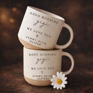 Op de afbeelding: Twee gestapelde crèmekleurige keramische mokken met een bruine basis. Elke mok heeft de zwarte tekst "GOOD MORNING gigi, WE LOVE YOU, SAMMY, ELLIE, OLIVER". Een witte madelief met een gele kern ligt bij de onderste mok.