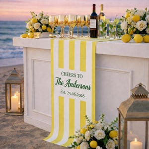 Puede incluir: Un bar en la playa decorado para una celebración, con un camino de mesa a rayas amarillas y blancas que dice "Cheers to The Andersons" y la fecha "Est. 25.06.2026". El bar está adornado con limones, flores, copas de vino y faroles.