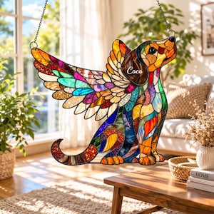 以下が含まれることがあります： カラフルなステンドグラスのアートワーク。犬が天使の羽を持ち、オレンジ、青、緑などの鮮やかなモザイク模様。犬の名前「Coco」が見える。チェーンで吊り下げられ、窓や壁に飾るのに適した装飾品です。