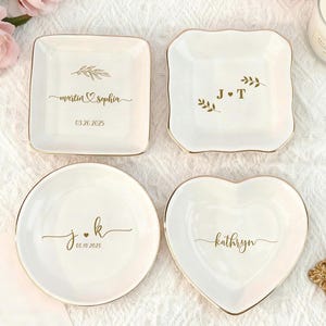 May include: Four white ceramic ring dishes with gold rims. One square dish has "martin & sophia" and a date. Another square dish has initials "J T". A round dish has initials "j.k" and a date. A heart-shaped dish has a name.