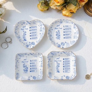 Puede incluir: Cuatro platos de cerámica blanca con motivos florales y texto en azul. Los platos tienen formas de corazón, redonda y cuadrada, cada uno con la frase 'God Says I Am' seguida de un nombre y una lista vertical de palabras descriptivas en tinta azul.