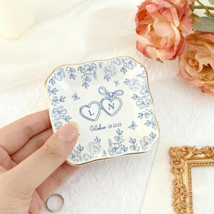 Puede incluir: Un peque&ntilde;o plato cuadrado de cer&aacute;mica blanca con borde dorado, con dise&ntilde;os florales azules y dos medallones en forma de coraz&oacute;n con las letras "L" y "N". La fecha "28 de octubre de 2025" tambi&eacute;n est&aacute; impresa en el plato.