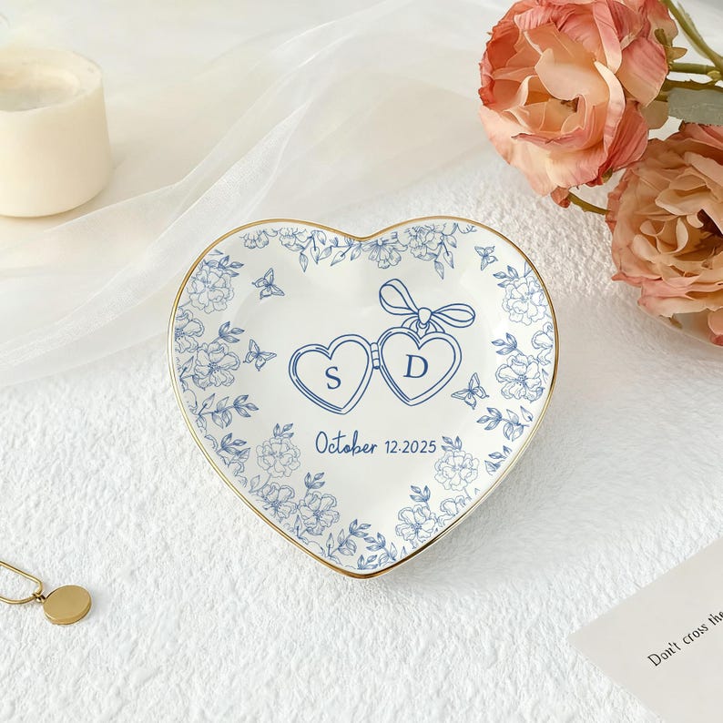 Puede incluir: Plato de cer&aacute;mica en forma de coraz&oacute;n con borde dorado, con dise&ntilde;os florales azules y dos corazones conectados con las iniciales "S" y "D". La fecha "12 de octubre de 2025" est&aacute; impresa debajo. Una pieza decorativa.