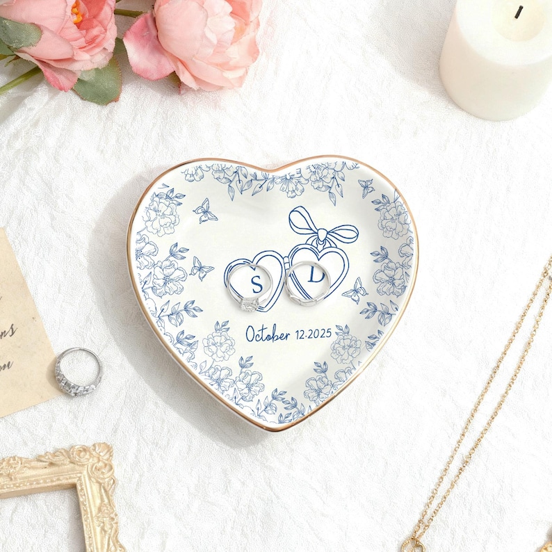 Puede incluir: Plato de cer&aacute;mica en forma de coraz&oacute;n con dise&ntilde;o floral azul y borde dorado. El plato contiene dos anillos, cada uno dentro de un coraz&oacute;n con las iniciales "S" y "D". La fecha "October 12-2025" est&aacute; impresa debajo.