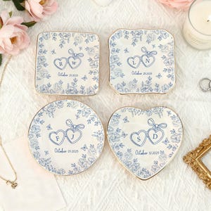Puede incluir: Cuatro posavasos de cer&aacute;mica blanca con dise&ntilde;os florales azules y bordes dorados. Cada posavasos presenta iniciales dentro de corazones y una fecha: 25 de octubre de 2025, 28 de octubre de 2025, 01 de octubre de 2025 y 12 de octubre de 2025.