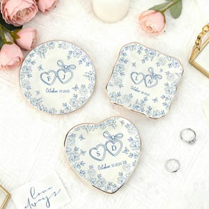 Puede incluir: Tres platos decorativos de cer&aacute;mica con dise&ntilde;os florales azules y bordes dorados. Cada plato presenta iniciales dentro de formas de coraz&oacute;n y una fecha. Uno es redondo, otro cuadrado y otro en forma de coraz&oacute;n. Tema de boda o aniversario.