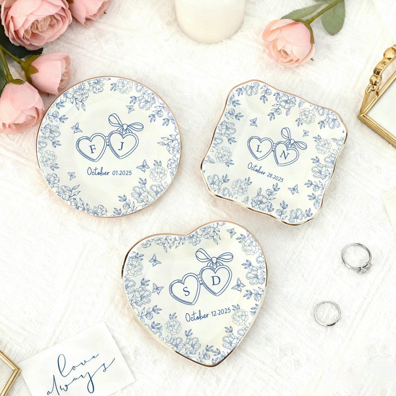 Puede incluir: Tres platos decorativos de cer&aacute;mica con dise&ntilde;os florales azules y bordes dorados. Cada plato presenta iniciales dentro de formas de coraz&oacute;n y una fecha. Uno es redondo, otro cuadrado y otro en forma de coraz&oacute;n. Tema de boda o aniversario.