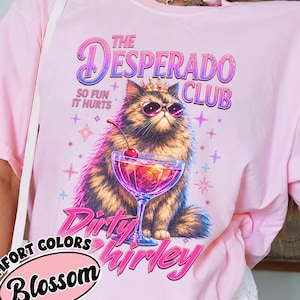 May include: A light pink t-shirt featuring a graphic of a fluffy cat wearing a crown and pink sunglasses, holding a cocktail with a cherry. The text reads THE DESPERADO CLUB, SO FUN IT HURTS, and Dirty Shirley. A Comfort Colors Blossom label is visible.