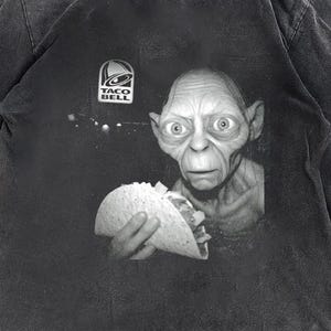 May include: Dark gray t-shirt featuring a black and white image of a character holding a taco. The Taco Bell logo is above the character's head. The shirt has a vintage, distressed look.