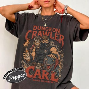 May include: A charcoal gray t-shirt featuring a graphic print with the text "DUNGEON CRAWLER CARL" and "YOU WILL NOT BREAK ME". The design depicts a masked character, a cat wearing a crown, a dinosaur, and a bottle labeled "JUG O' BOOM".