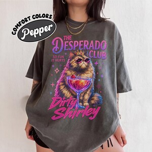 May include: A charcoal gray t-shirt featuring a graphic of a fluffy cat wearing a crown and sunglasses next to a cocktail. The text reads The Desperado Club, So Fun It Hurts, and Dirty Shirley. A Comfort Colors Pepper brand logo is in the corner.