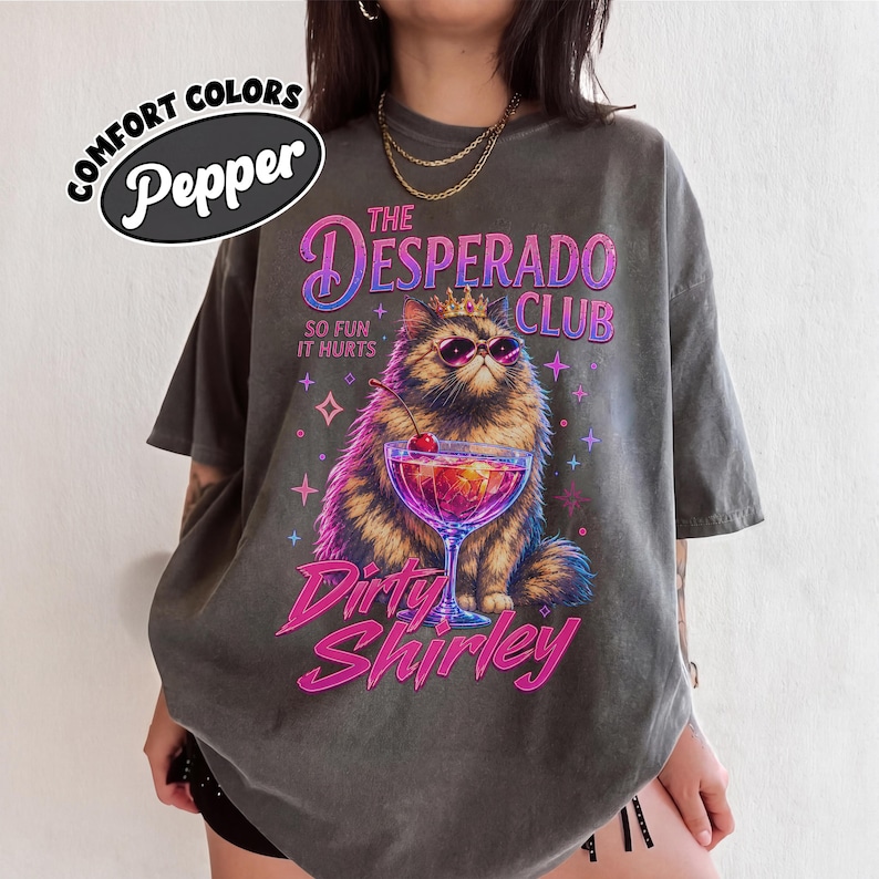 May include: A charcoal gray t-shirt featuring a graphic of a fluffy cat wearing a crown and sunglasses next to a cocktail. The text reads The Desperado Club, So Fun It Hurts, and Dirty Shirley. A Comfort Colors Pepper brand logo is in the corner.