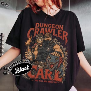 May include: A black t-shirt featuring a graphic print of a warrior, a cat wearing a crown, and a dinosaur. The text reads "DUNGEON CRAWLER", "JUG O' BOOM", "CARL", and "YOU WILL NOT BREAK ME". A circular label on the left side displays "COMFORT COLORS Black".