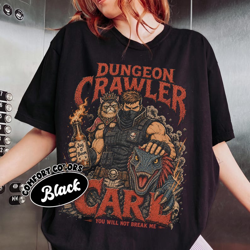 May include: A black t-shirt featuring a graphic print of a warrior, a cat wearing a crown, and a dinosaur. The text reads "DUNGEON CRAWLER", "JUG O' BOOM", "CARL", and "YOU WILL NOT BREAK ME". A circular label on the left side displays "COMFORT COLORS Black".