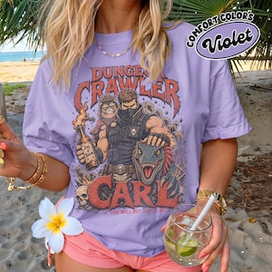May include: A violet t-shirt featuring a graphic print with the text "DUNGEON CRAWLER CARL" and "YOU WILL NOT BREAK ME". The design depicts a masked character, a cat wearing a crown, a dinosaur, and a bottle labeled "Jug O Boom" with skulls.