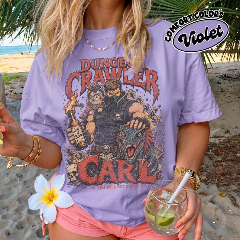 May include: A violet t-shirt featuring a graphic print with the text "DUNGEON CRAWLER CARL" and "YOU WILL NOT BREAK ME". The design depicts a masked character, a cat wearing a crown, a dinosaur, and a bottle labeled "Jug O Boom" with skulls.