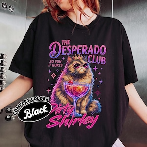 May include: A black t-shirt featuring a graphic of a fluffy cat wearing a gold crown and pink sunglasses. The cat sits behind a cocktail glass with a cherry. Text reads: THE DESPERADO CLUB, SO FUN IT HURTS, and Dirty Shirley in pink and purple.