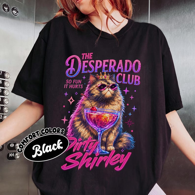 May include: A black t-shirt featuring a graphic of a fluffy cat wearing a gold crown and pink sunglasses. The cat sits behind a cocktail glass with a cherry. Text reads: THE DESPERADO CLUB, SO FUN IT HURTS, and Dirty Shirley in pink and purple.