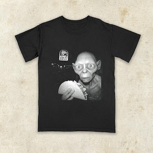 K&ouml;nnte beinhalten: Schwarzes T-Shirt mit einem Schwarz-Wei&szlig;-Bild einer Figur, die einen Taco h&auml;lt, mit dem Taco Bell-Logo. Das Grafik-T-Shirt ist eine humorvolle Parodie.