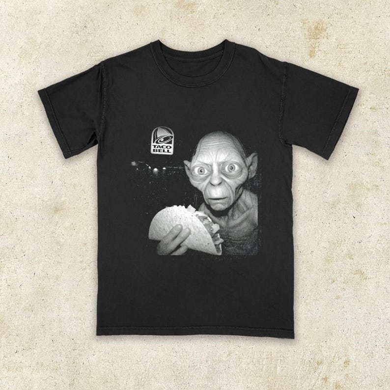 K&ouml;nnte beinhalten: Schwarzes T-Shirt mit einem Schwarz-Wei&szlig;-Bild einer Figur, die einen Taco h&auml;lt, mit dem Taco Bell-Logo. Das Grafik-T-Shirt ist eine humorvolle Parodie.
