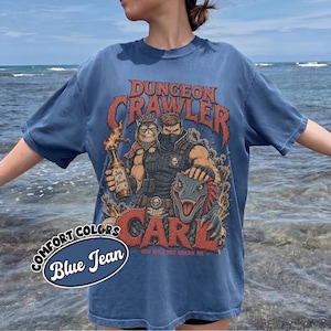 May include: A blue jean colored t-shirt featuring a graphic print with the text DUNGEON CRAWLER CARL. The design depicts a masked character, a cat, and a dinosaur, with the character holding a bottle labeled JUG O BOOM and the text YOU WILL NOT BREAK ME.