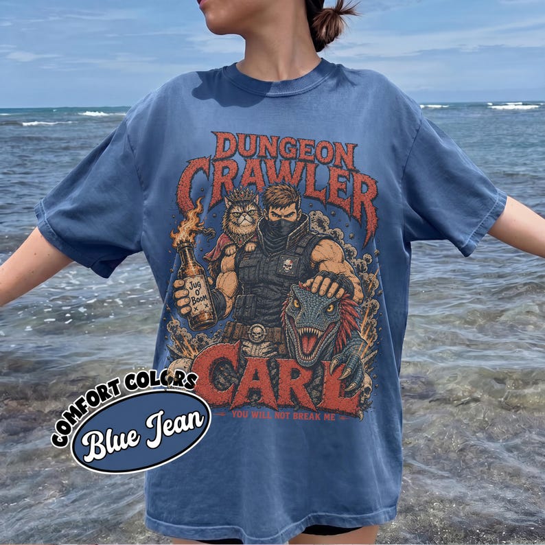 May include: A blue jean colored t-shirt featuring a graphic print with the text DUNGEON CRAWLER CARL. The design depicts a masked character, a cat, and a dinosaur, with the character holding a bottle labeled JUG O BOOM and the text YOU WILL NOT BREAK ME.