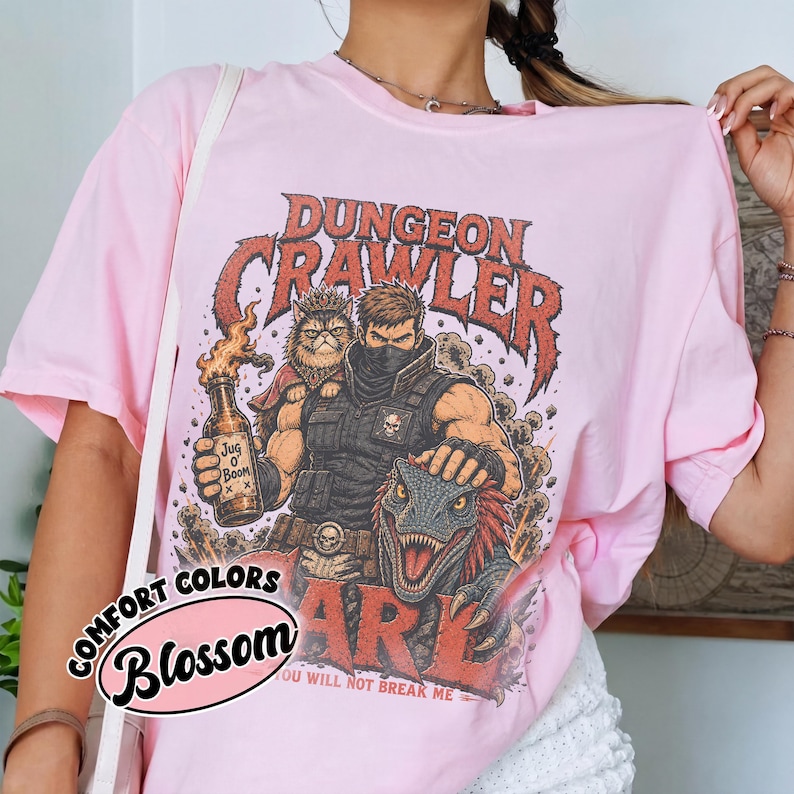 May include: A light pink t-shirt featuring a graphic print of a masked warrior, a cat, and a dinosaur. The text reads Dungeon Crawler, Jug O' Boom, and You Will Not Break Me. A Comfort Colors Blossom logo is visible in the lower left corner of the image.