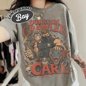 May include: A grey t-shirt featuring a graphic print with the text "Dungeon Crawler Carl" and "Jug O Boom". The illustration depicts a masked character, a cat wearing a crown, and a dinosaur. The shirt includes a "Comfort Colors Bay" label.