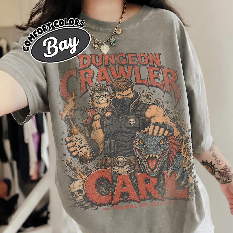 May include: A grey t-shirt featuring a graphic print with the text "Dungeon Crawler Carl" and "Jug O Boom". The illustration depicts a masked character, a cat wearing a crown, and a dinosaur. The shirt includes a "Comfort Colors Bay" label.