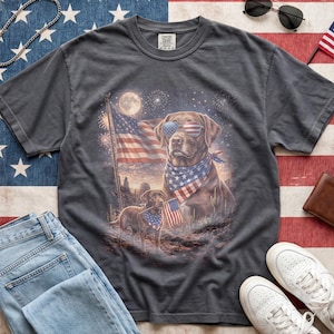 May include: Dark gray t-shirt featuring a patriotic dog graphic. The design includes a dog wearing sunglasses and a bandana with an American flag, fireworks, and a full moon. The shirt is paired with blue jeans and white sneakers.