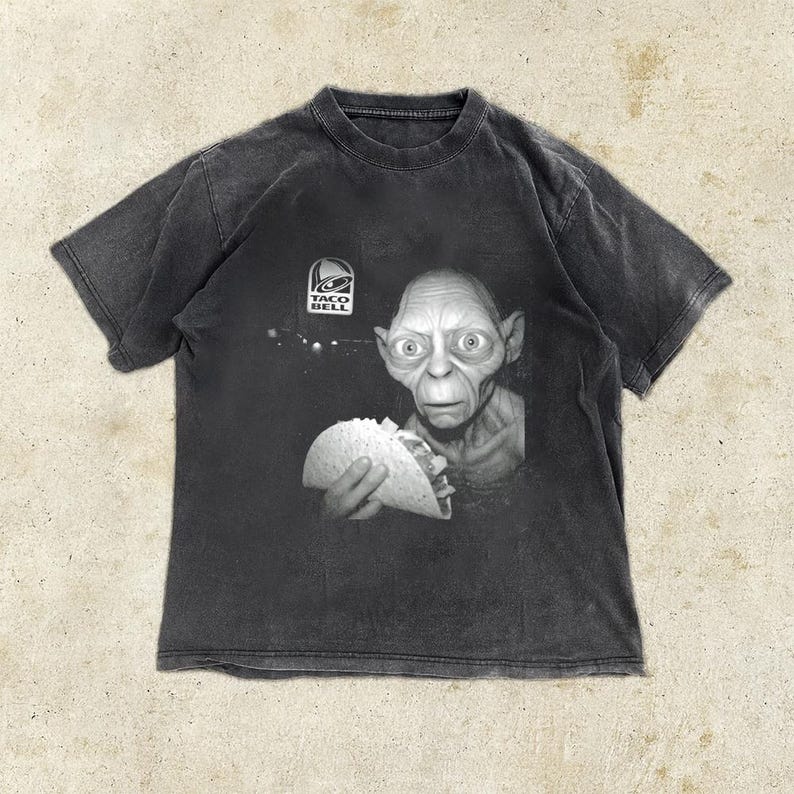 May include: Dark gray t-shirt featuring a black and white image of a character holding a taco. The Taco Bell logo is above the character's head. The shirt has a vintage, distressed look.