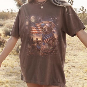 May include: Brown t-shirt featuring a patriotic design with two Labrador Retrievers. One dog wears sunglasses and a bandana with an American flag pattern. The other dog holds a small American flag. Fireworks and the American flag are in the background.