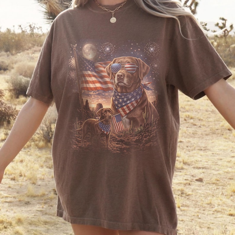May include: Brown t-shirt featuring a patriotic design with two Labrador Retrievers. One dog wears sunglasses and a bandana with an American flag pattern. The other dog holds a small American flag. Fireworks and the American flag are in the background.